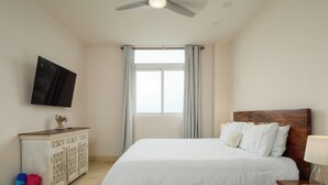 A bedroom with a wooden bed, a TV mounted on the wall, a window with curtains, and a wooden nightstand with a lamp. - Coastal Retreat 2BR Skyline Marina View (Río Hato)