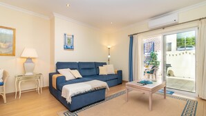 A living room with a blue sofa, a small table, a lamp, and a view of the outside through a glass door. - Casa Peggy - Communal pool & garden, just 500m from the beach (Carvoeiro)