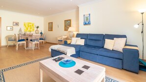 A living room with a blue sofa, a coffee table, and a dining area with chairs and a table. - Casa Peggy - Communal pool & garden, just 500m from the beach (Carvoeiro)
