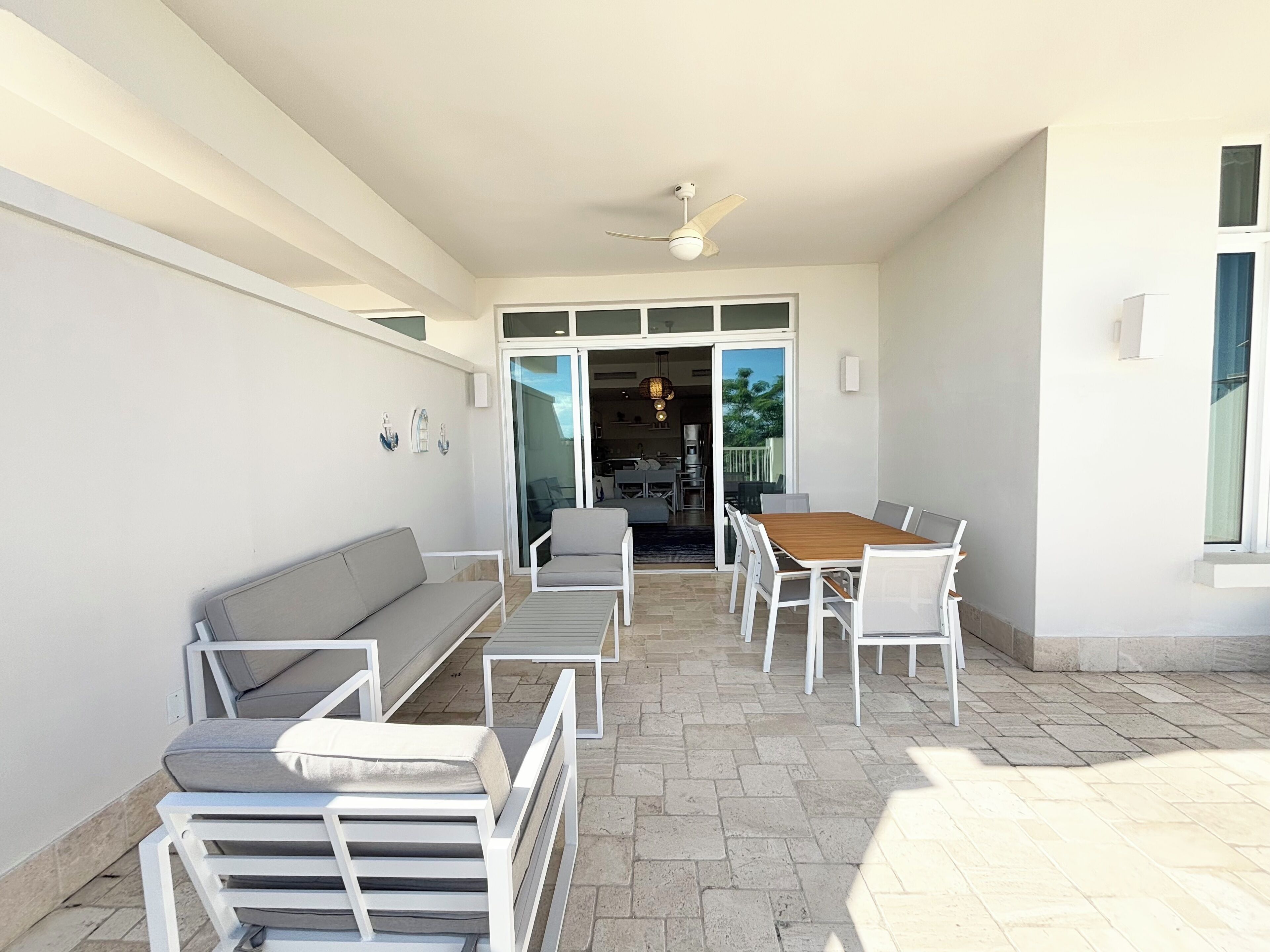 Eine sonnendurchflutete Terrasse mit Holztisch und weißen Stühlen, einem grauen Sofa und einem Deckenventilator.