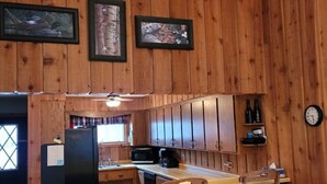 A wooden cabin interior with a kitchen area featuring a bar with stools, a ceiling fan, and framed artwork on the walls. - Peaceful Cavalier Townhome #2 Near Lake Owen (Cable)