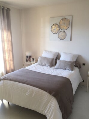 Uma cama bem arrumada com colcha branca e marrom, uma mesa de cabeceira com abajur e uma obra de arte na parede acima da cama.