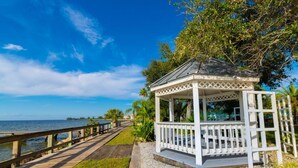 A gazebo with a white lattice roof and railing, situated on a gravel path beside a wooden boardwalk leading to a body of water.
