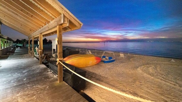 A wooden pier extending over a beach with a sunset view, a covered seating area, and a row of beach chairs.