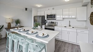 Une cuisine moderne avec des meubles blancs, un îlot central doté de tabourets de bar, des appareils en acier inoxydable et une table à manger pour quatre personnes.