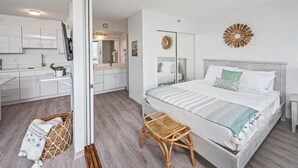Une chambre à coucher moderne avec un grand lit, une table de chevet, un fauteuil en osier et une salle de bain visible à travers une porte ouverte.