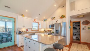 A modern kitchen with white cabinets, a granite countertop, a stainless steel refrigerator, and a large island with bar stools. - The Pup Deck - Duck, Carolina Dunes, 420 Yds to Beach, Pool, Hot Tub, Pets OK, Flex Stays (Duck)