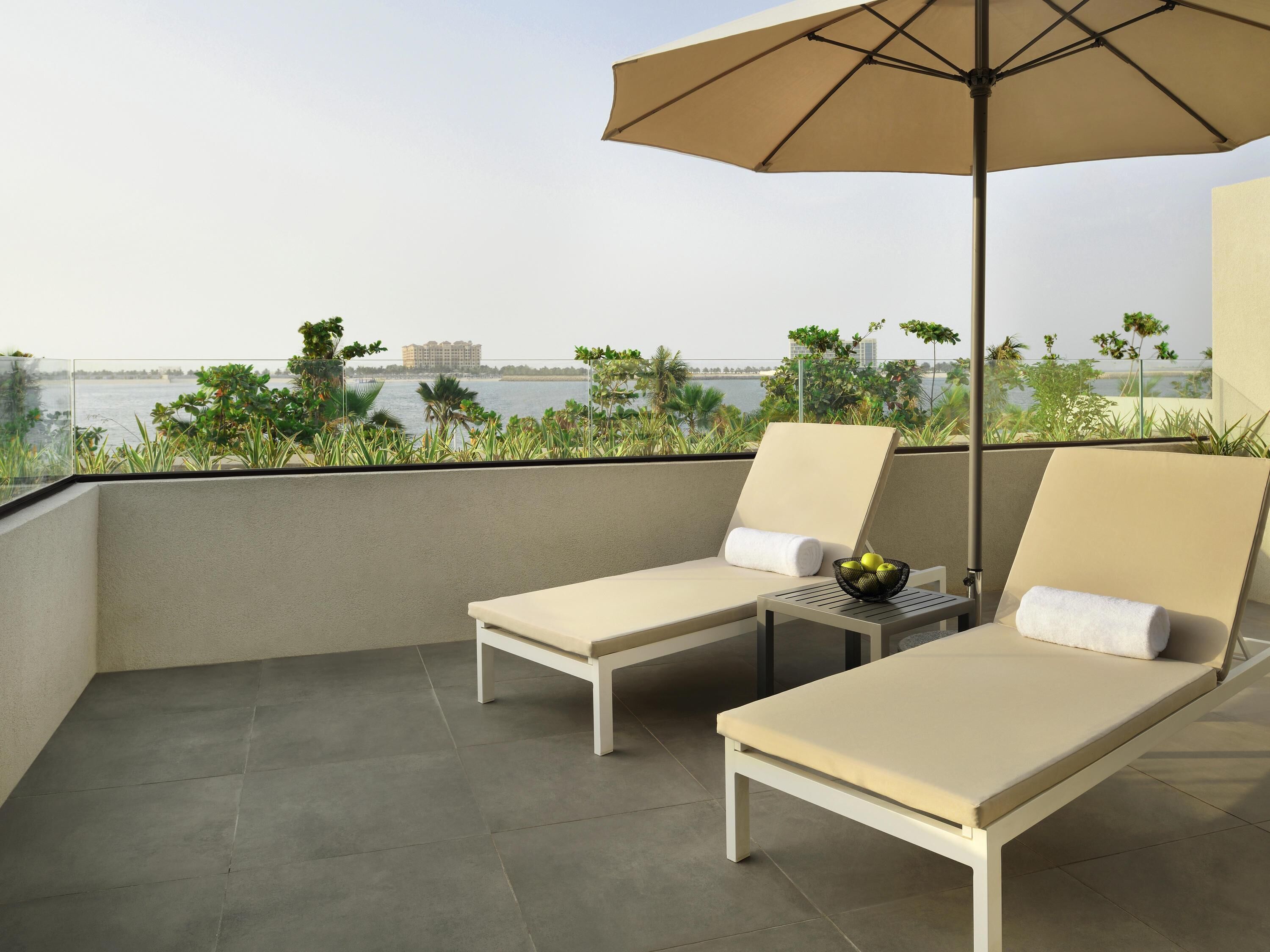 Ein Balkon mit zwei Liegestühlen, einem Tisch mit Früchten und Blick auf Wasser und Gebäude.