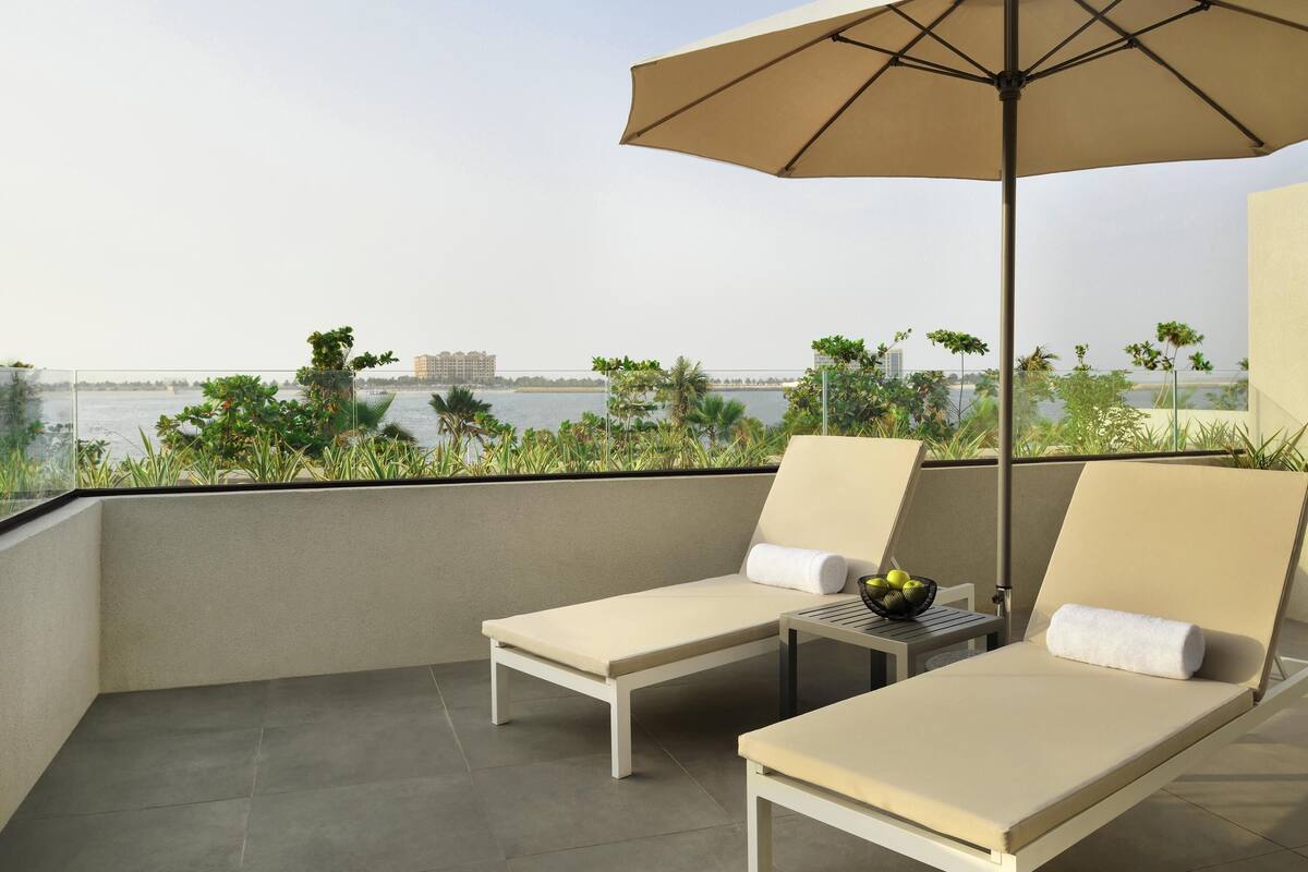 Ein Balkon mit zwei Liegestühlen, einem Tisch mit Früchten und Blick auf Wasser und Gebäude.