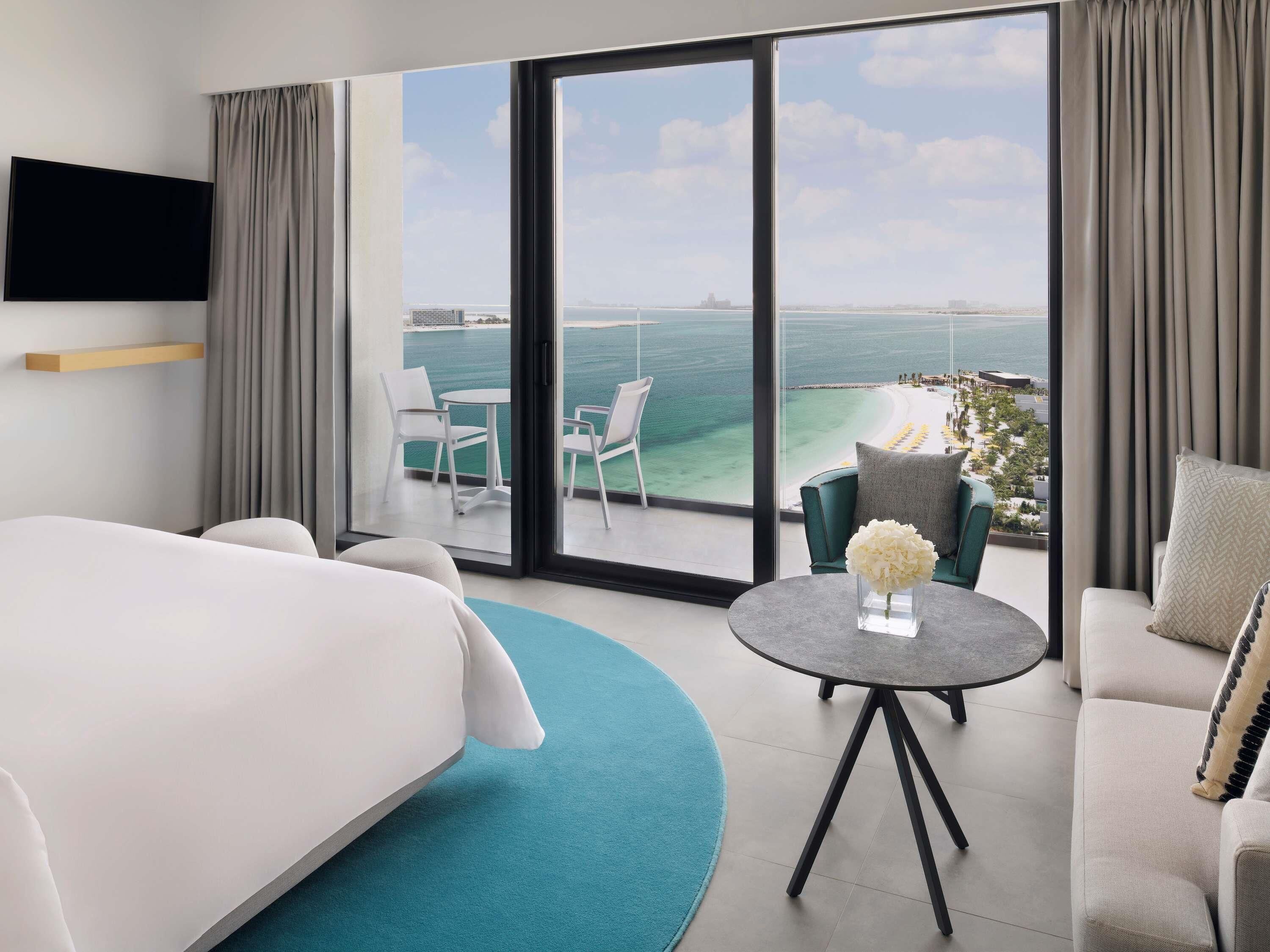 Een hotelkamer met een bed, een ronde tafel met een vaas, een bank en een balkon met uitzicht op zee.