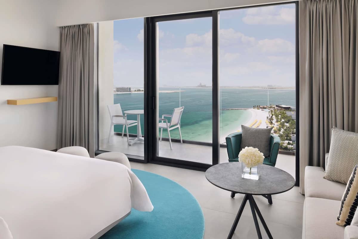 A hotel room with a bed, a round table with a vase, a sofa, and a balcony with a view of the sea.