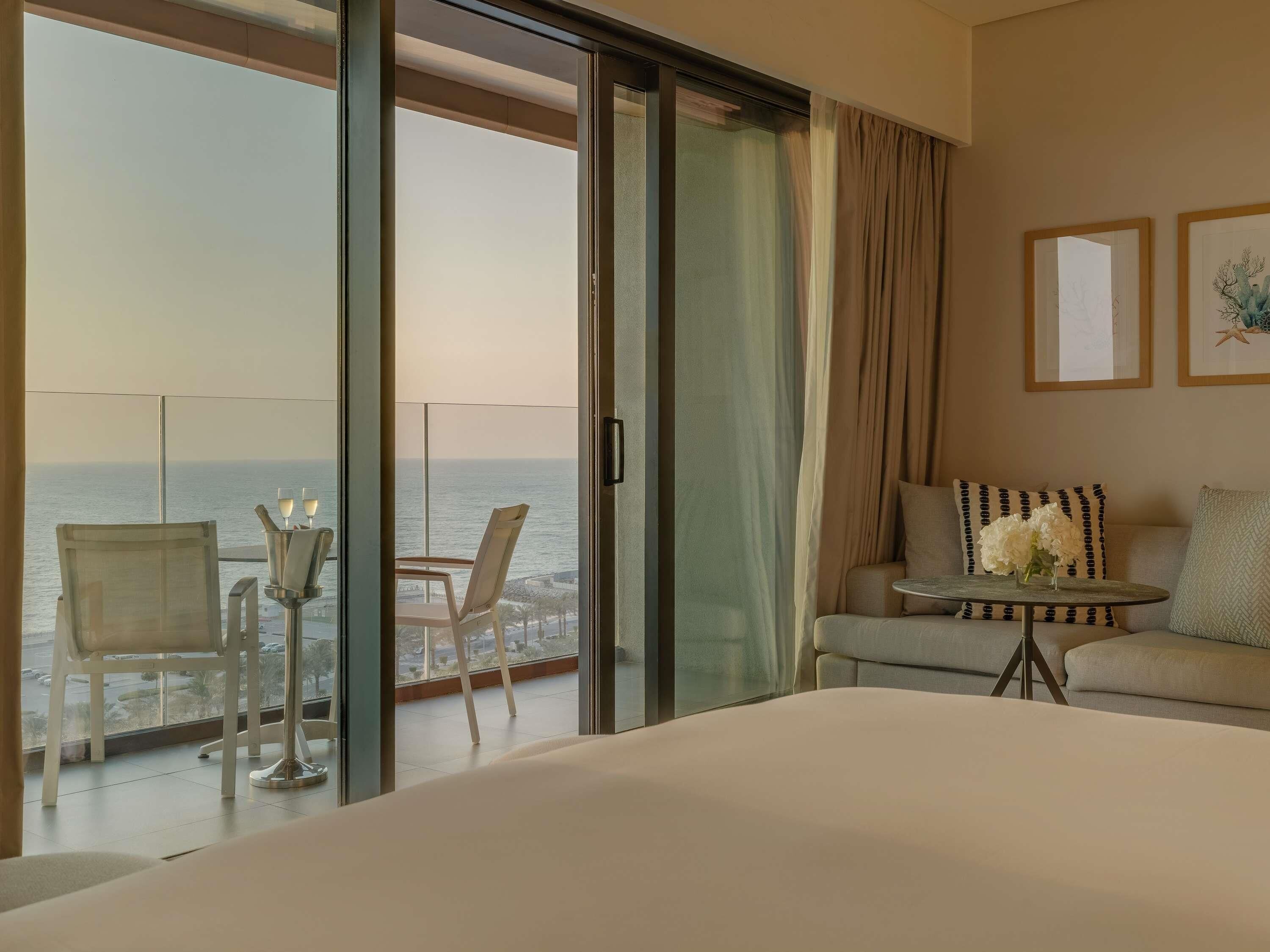Habitación de hotel con balcón, cama, sofá y una pequeña mesa con un jarrón de flores.