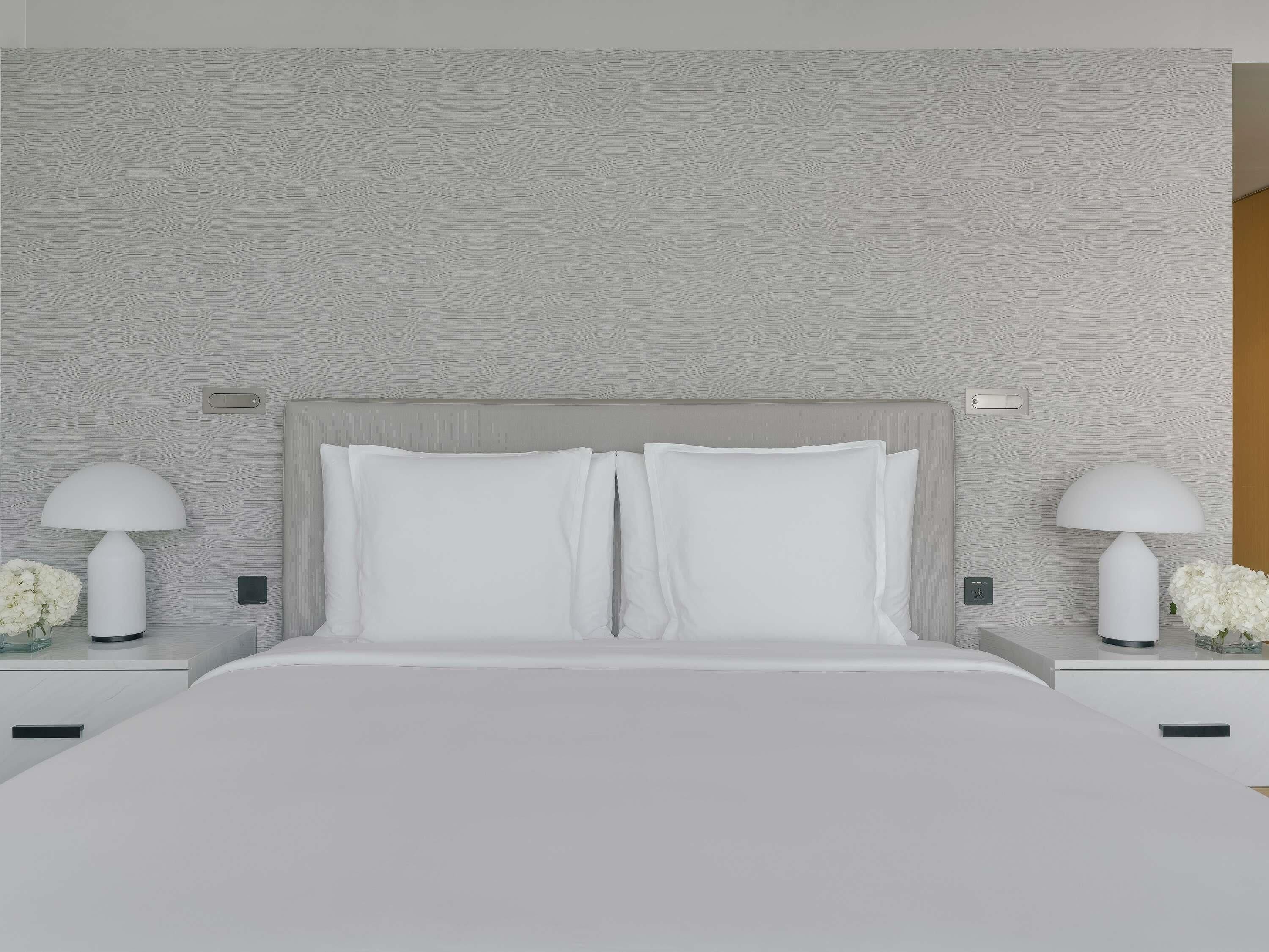 Une chambre à coucher moderne avec un mur texturé de couleur grise, un grand lit avec une literie blanche, deux tables de chevet avec des lampes et un petit vase avec des fleurs.