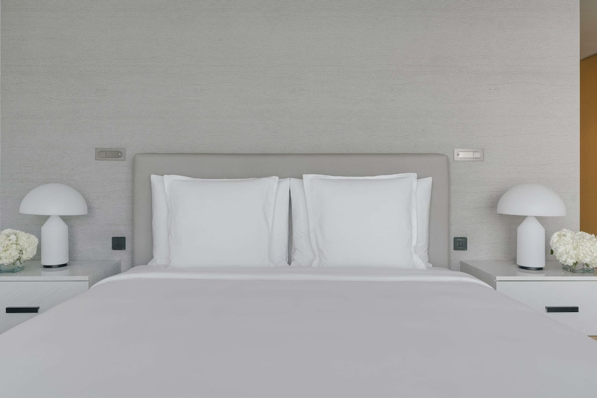 A modern bedroom with a grey textured wall, a large bed with white bedding, two bedside tables with lamps, and a small vase with flowers.