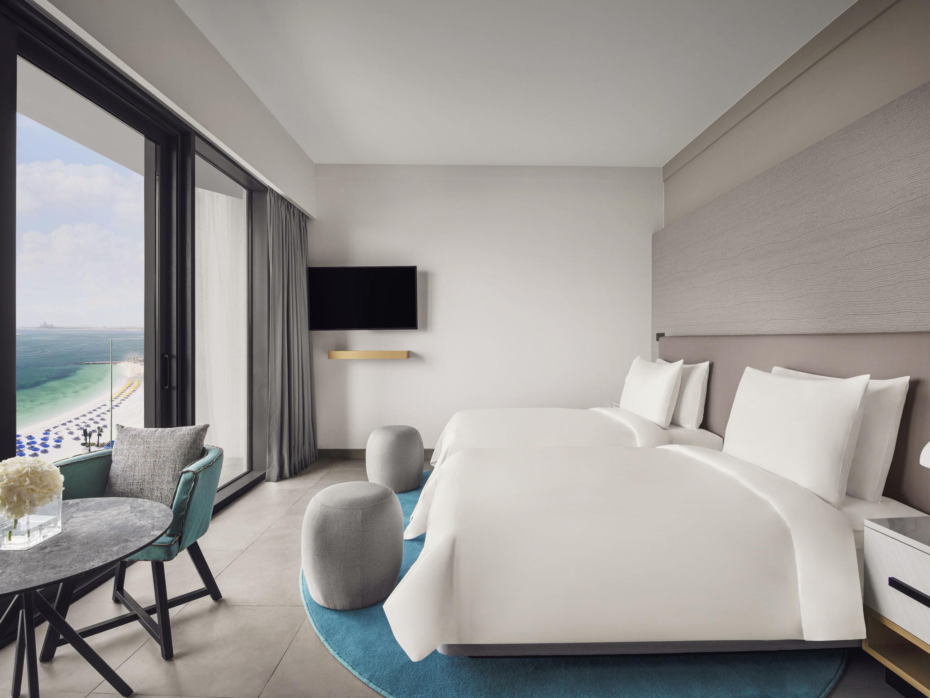 Een moderne hotelkamer met een groot bed, een aan de muur bevestigde televisie, een zithoek met een tafel en stoelen, en uitzicht op het strand.