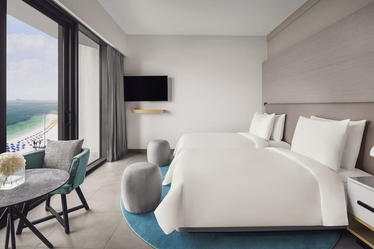 Ein modernes Hotelzimmer mit einem großen Bett, einem an der Wand montierten Fernseher, einem Essbereich mit Tisch und Stühlen sowie Blick auf den Strand.