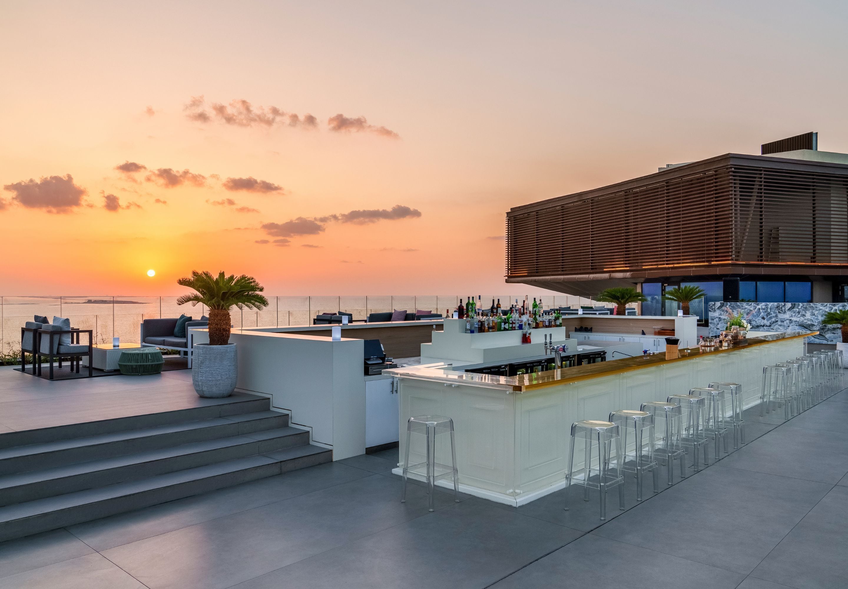 A rooftop bar with a bar counter, seating, and a view of the sunset over the ocean.