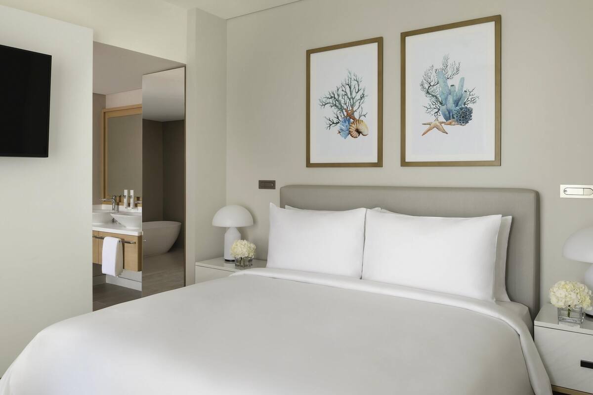 Ein modernes Hotelzimmer mit einem großen Bett, Nachttischen mit Lampen, einem Flachbildfernseher und zwei gerahmten Kunstwerken an der Wand.
