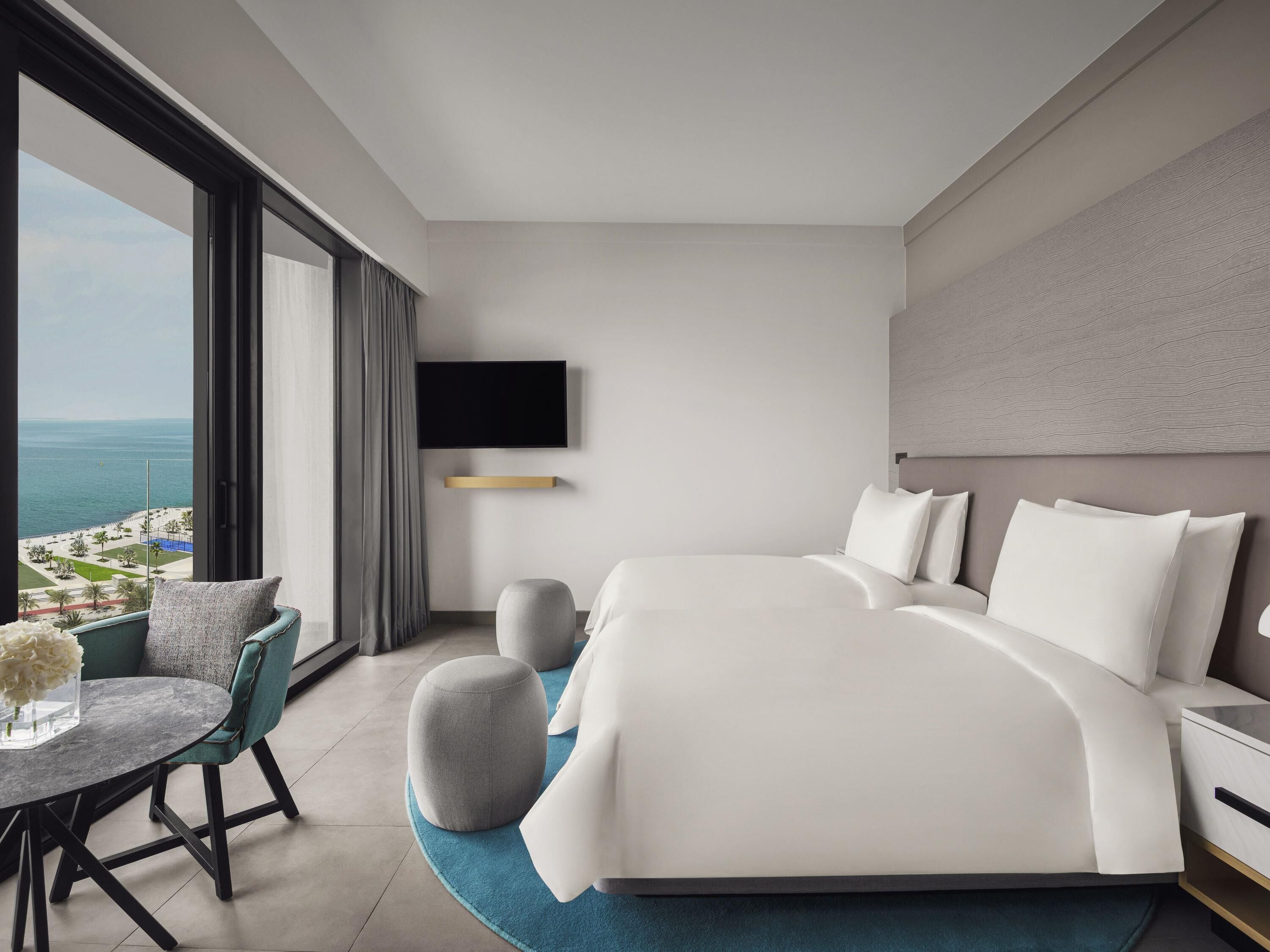 Een moderne hotelkamer met een groot bed, een flatscreen televisie aan de muur, een rond poefje, een klein rond tafeltje met een bloevas en uitzicht op het strand door grote ramen.