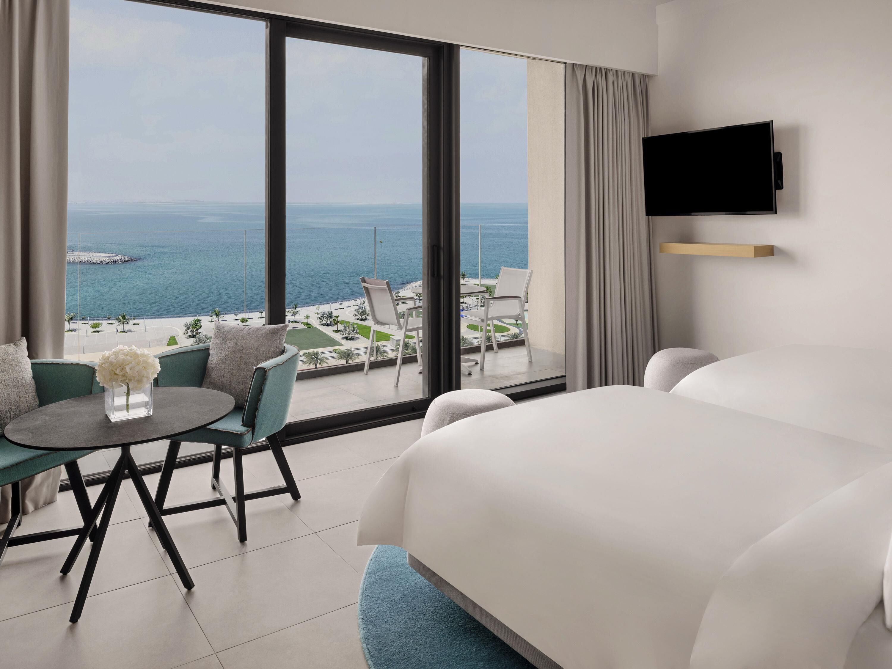 A hotel room with a large bed, a small round table with a vase of flowers, a sofa, and a view of the sea and a beach.