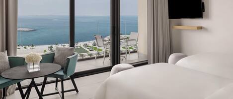 A hotel room with a large bed, a small round table with a vase of flowers, a sofa, and a view of the sea and a beach.