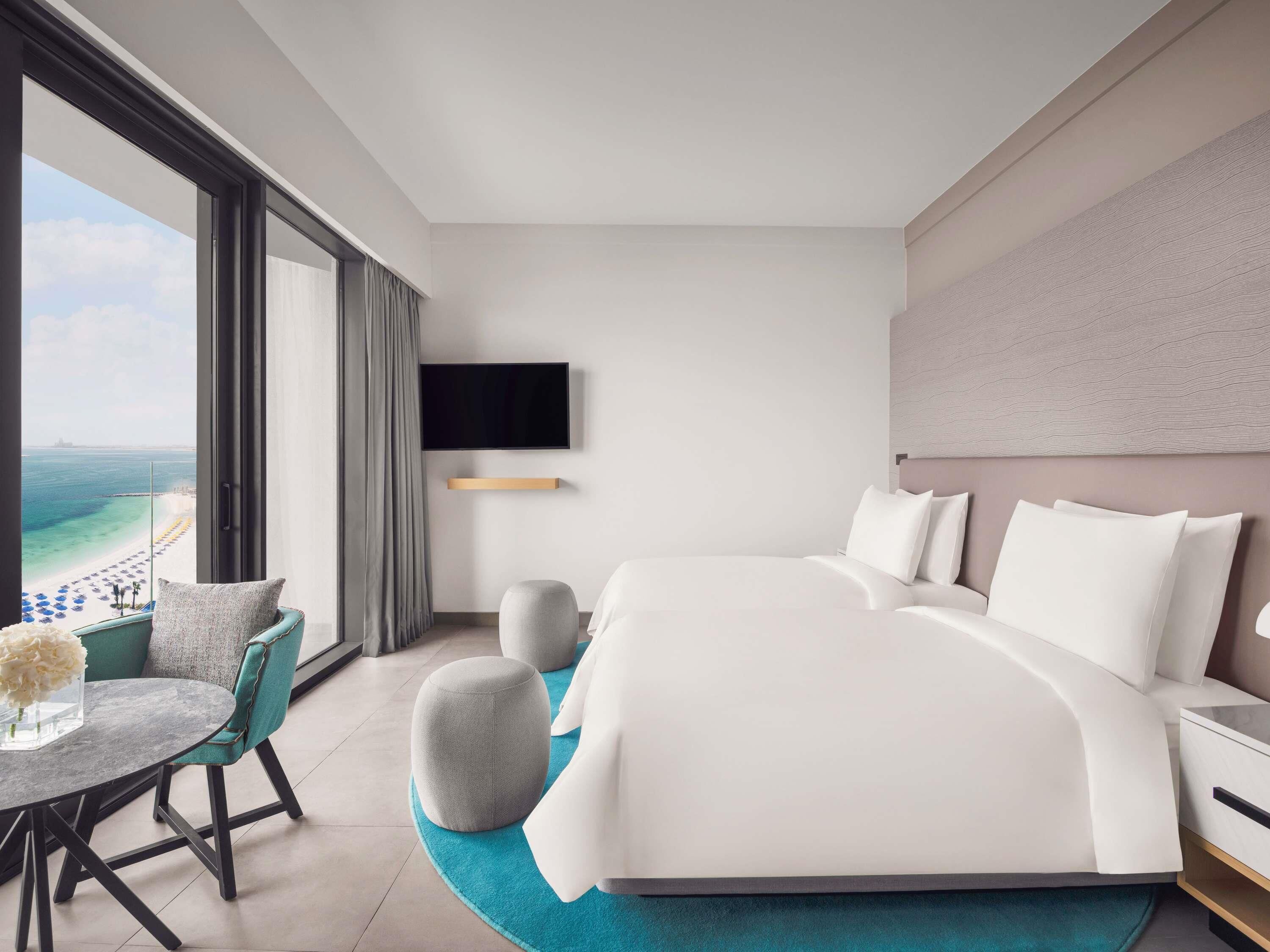 Een moderne hotelkamer met een groot bed, een aan de muur bevestigde televisie, een balkon met uitzicht op het strand en een zithoek met een tafel en stoelen.