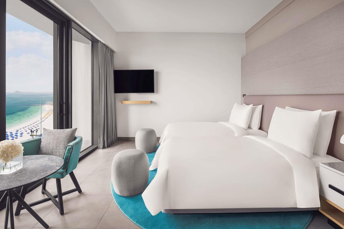 A modern hotel room with a large bed, a TV mounted on the wall, a balcony with a view of the beach, and a dining area with a table and chairs.