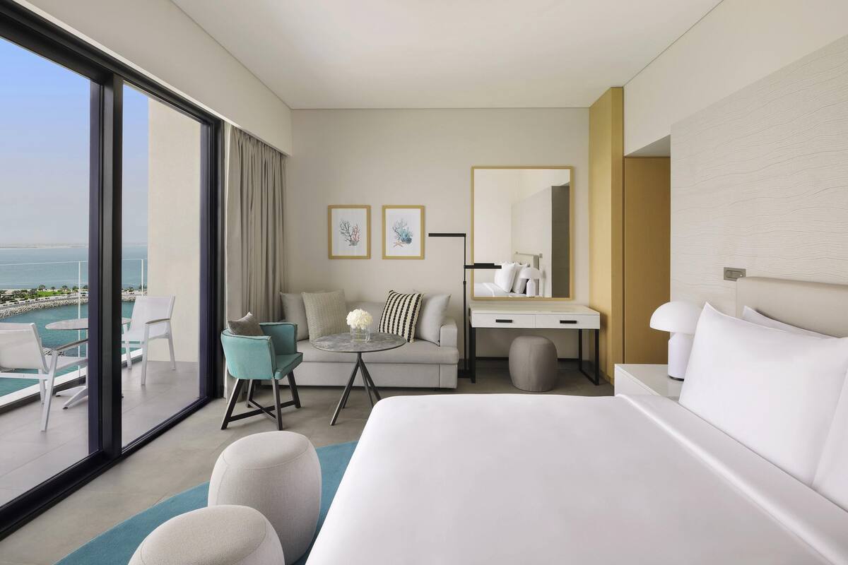 Ein modernes Hotelzimmer mit einem großen Bett, einer Sitzecke mit einer Couch und Sesseln, einem Schreibtisch mit Spiegel sowie Meerblick durch große Fenster.