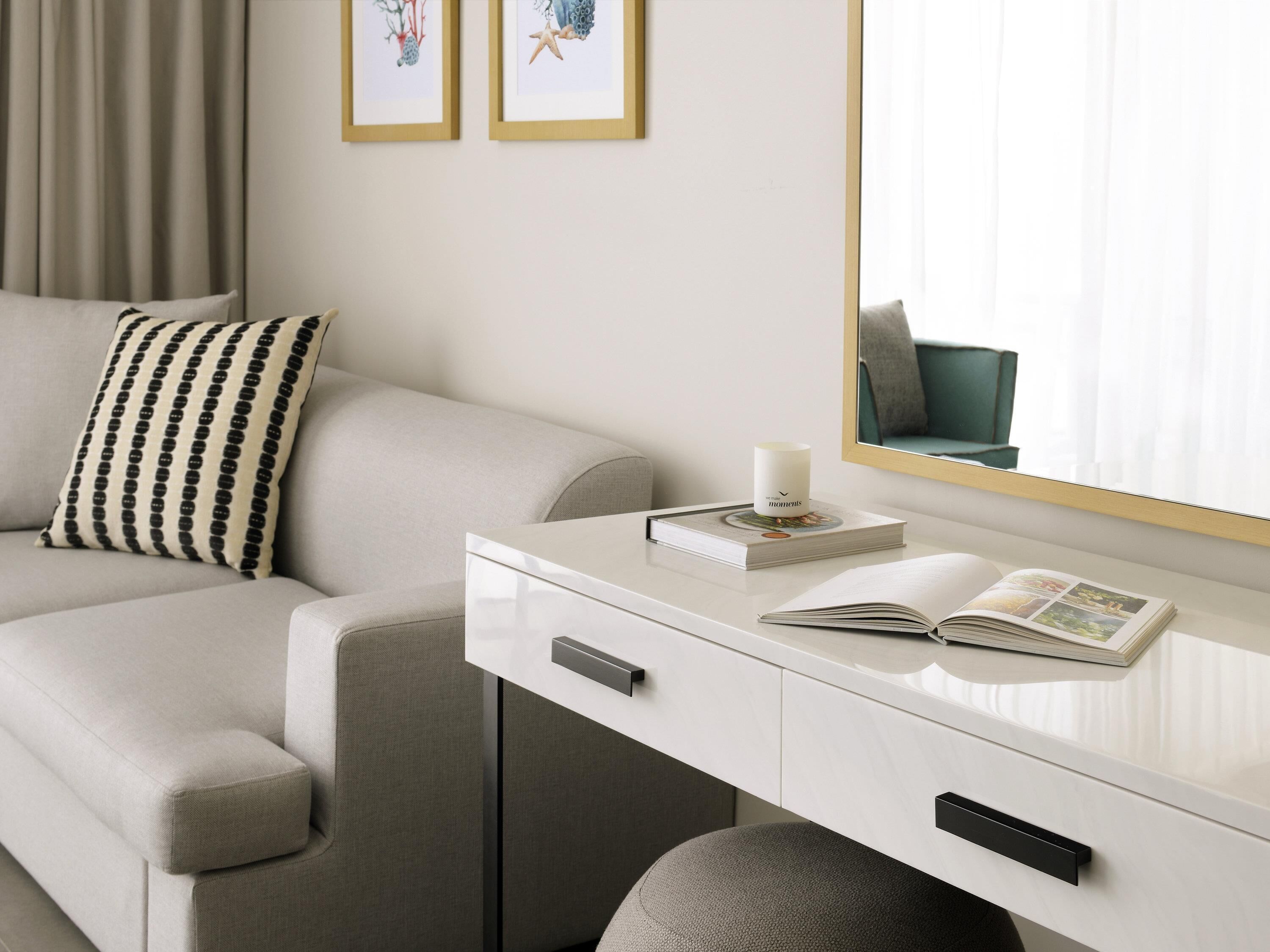 Une chambre d’hôtel moderne avec un canapé, une table de chevet sur laquelle se trouvent un livre et une bougie, et un miroir qui reflète la pièce.