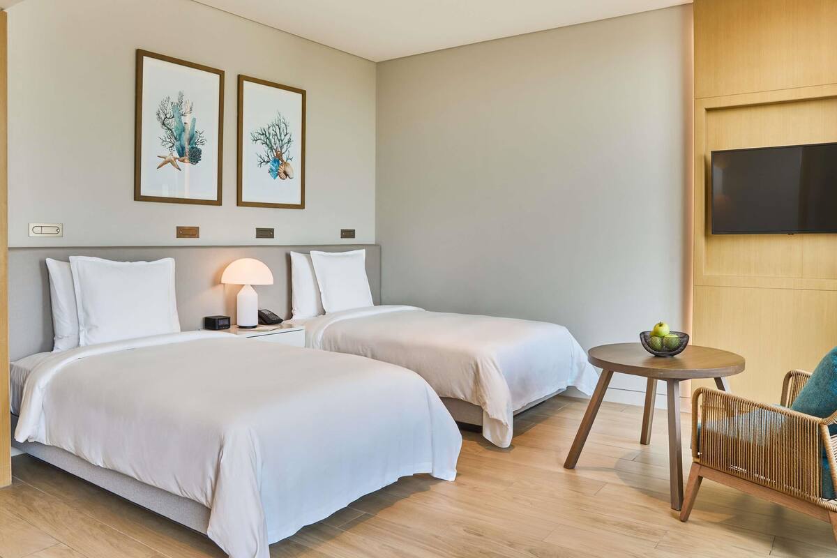 Ein Hotelzimmer mit zwei Betten, einem Nachttisch mit Lampe, einem kleinen runden Tisch mit Obstschale und einem an der Wand befestigten Fernseher.