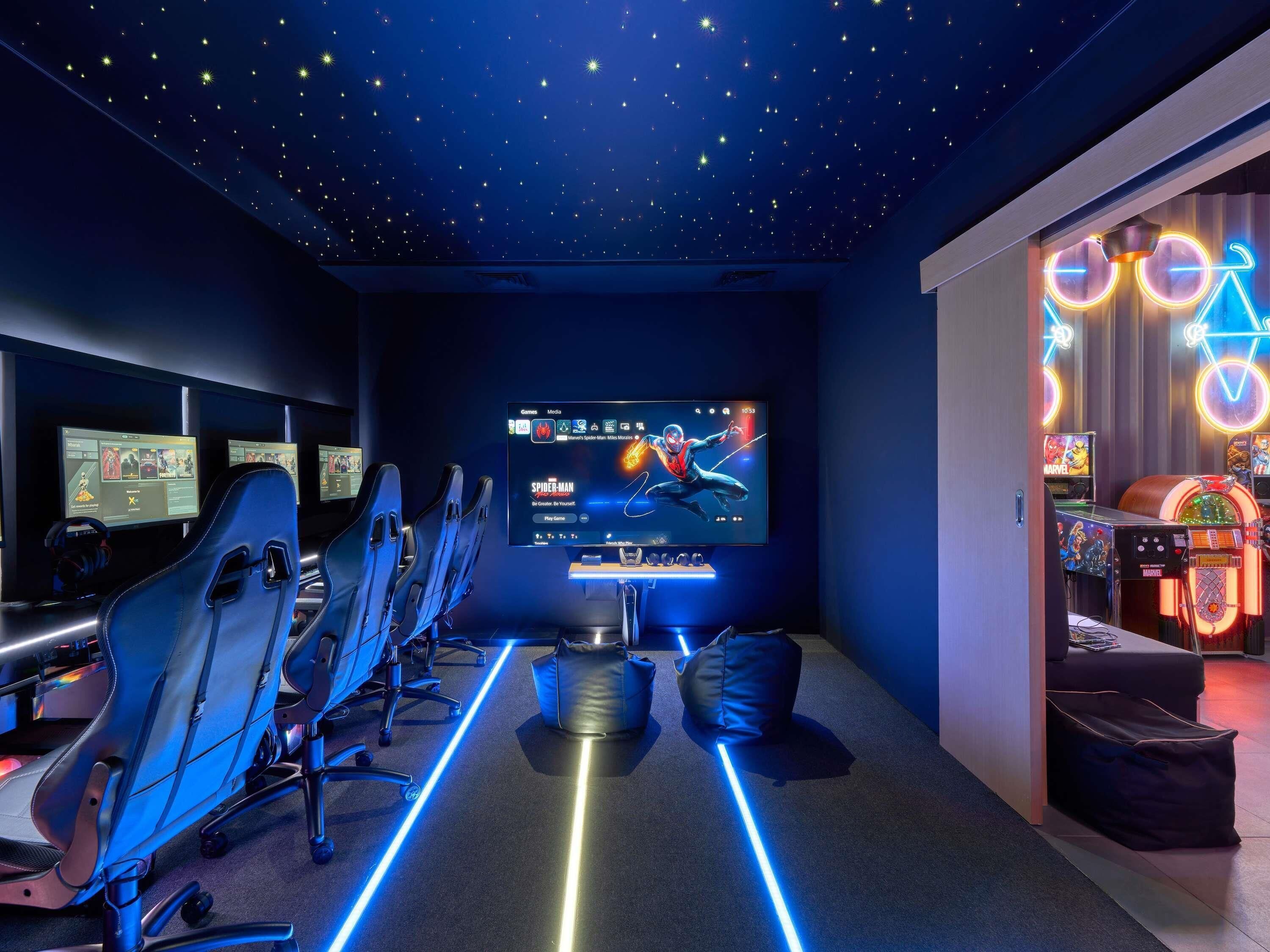 Ein Spielzimmer mit mehreren Gaming-Stationen, einem großen Bildschirm, der ein Spiel zeigt, und Neonbeleuchtung.