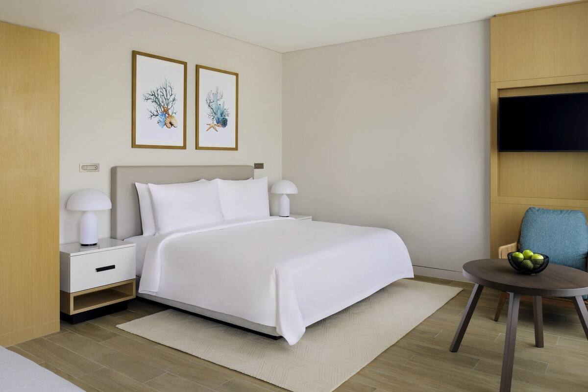 Ein modernes Hotelzimmer mit einem Bett, Nachttischen, einem runden Tisch mit Obst und Kunstwerken an der Wand.
