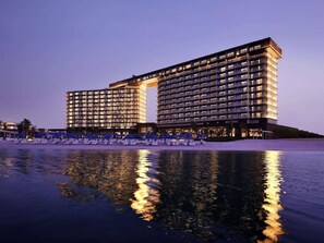 Een modern hotel met een glazen vijver ervoor, met een grote, verlichte ingang en meerdere balkons.