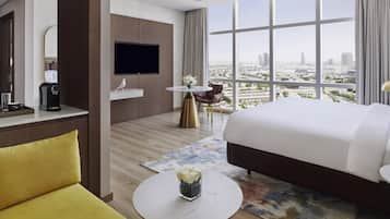 A modern hotel room with a large bed, a flat-screen TV, a coffee machine, a small table with a vase of flowers, and a view of the city through large windows.