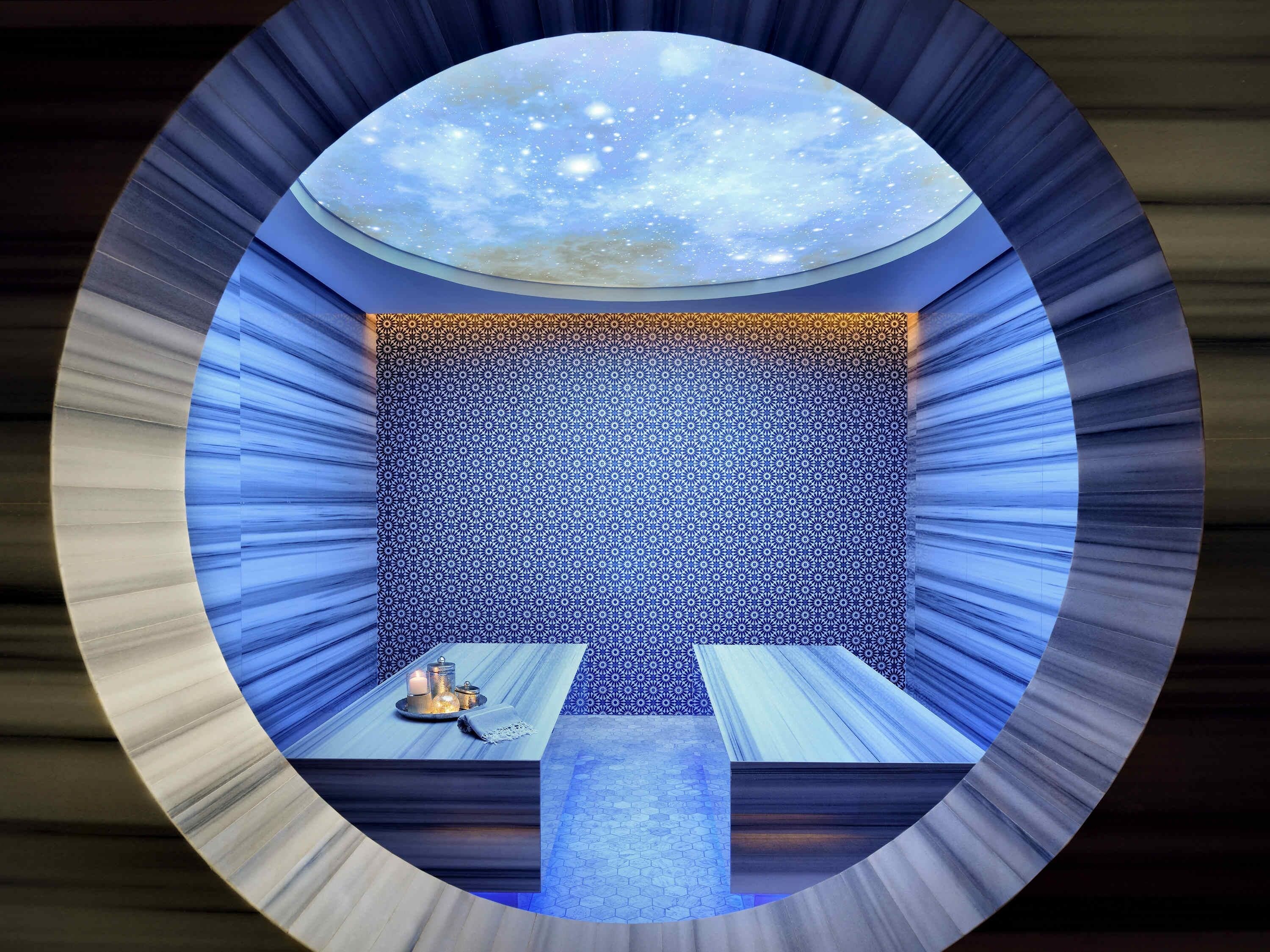 A room with a patterned wall, a table with candles, and a view of the sky through a circular opening.