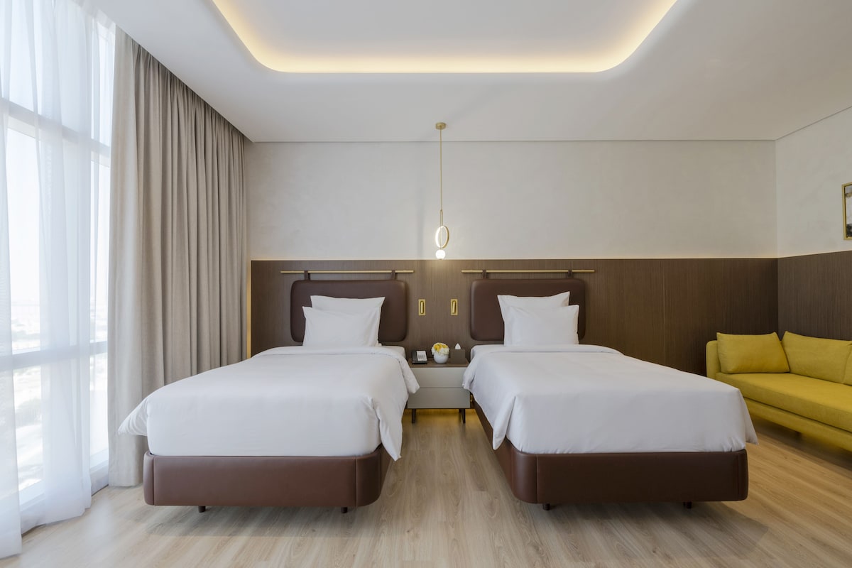 Une chambre d’hôtel avec deux lits, un canapé, une table de chevet et une grande fenêtre avec des rideaux.
