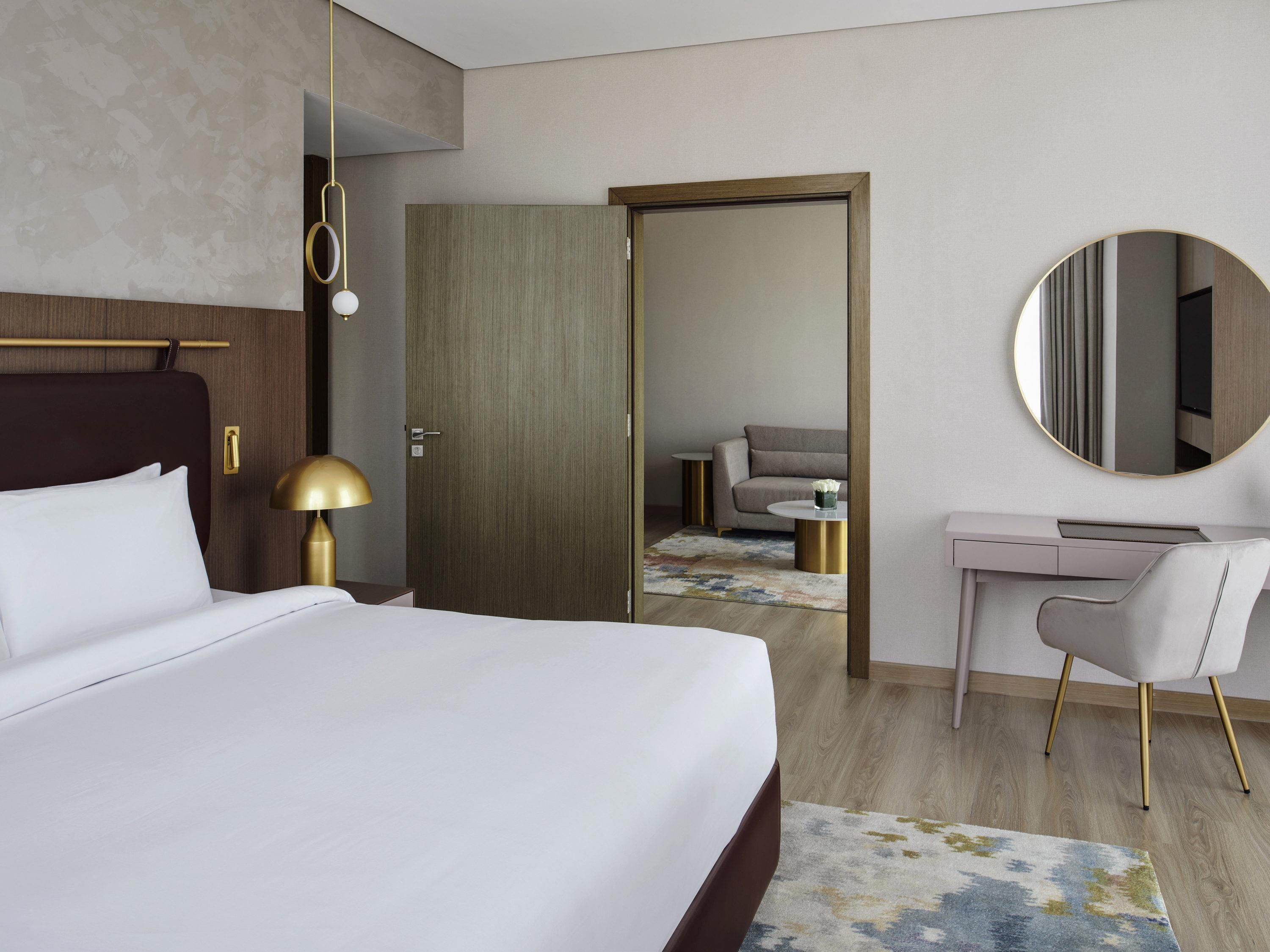 A modern hotel room with a large bed, a desk, a chair, a round mirror, and a wardrobe.