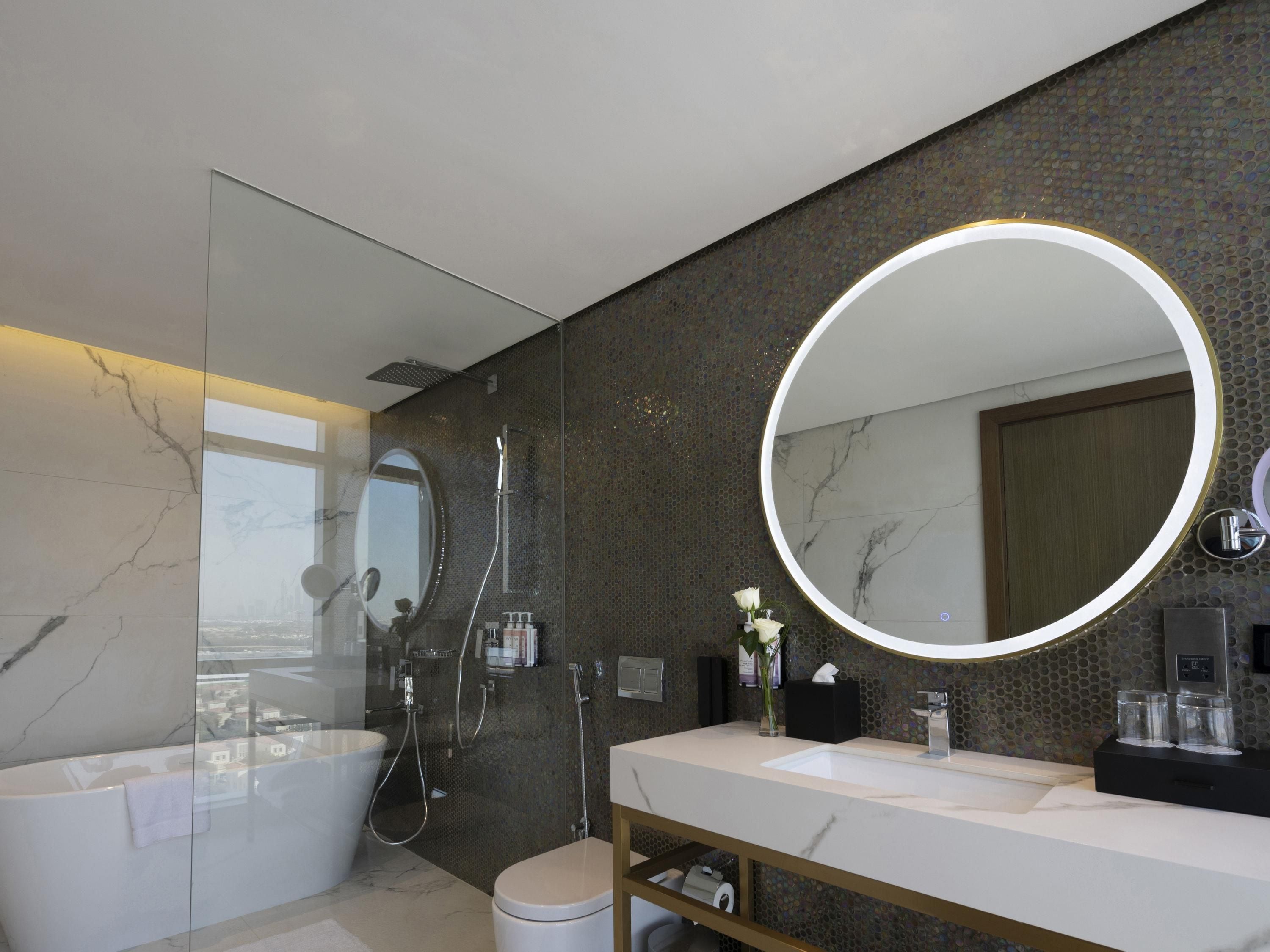 A modern bathroom with a glass-enclosed shower, a large round mirror, and a marble-tiled vanity.