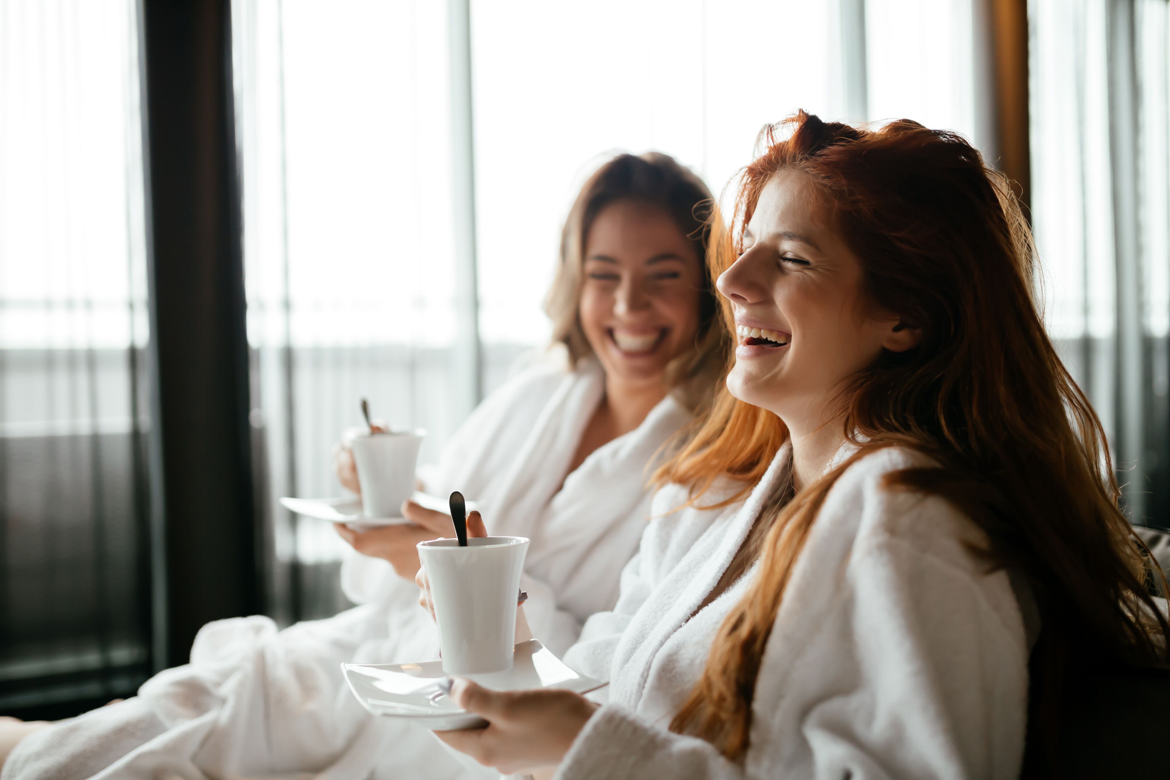 Two individuals in white robes sitting by a window, each holding a white cup with a spoon.