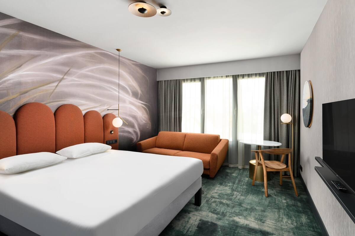 A modern hotel room with a large bed, a sofa, a small table, a chair, a TV, and a wall with a large abstract painting.