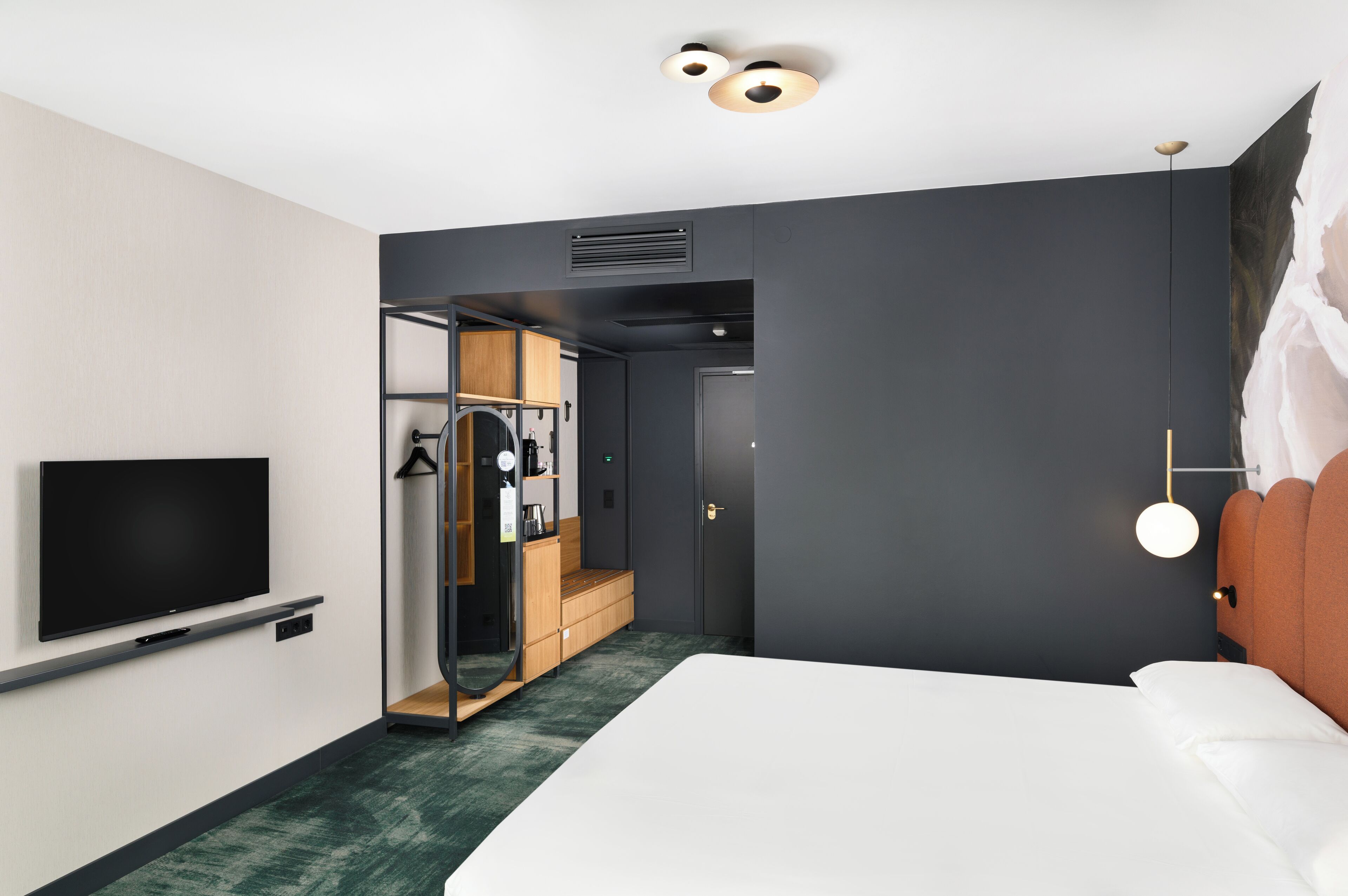 A modern hotel room with a large bed, a flat-screen TV mounted on the wall, a walk-in closet, and a dark accent wall.