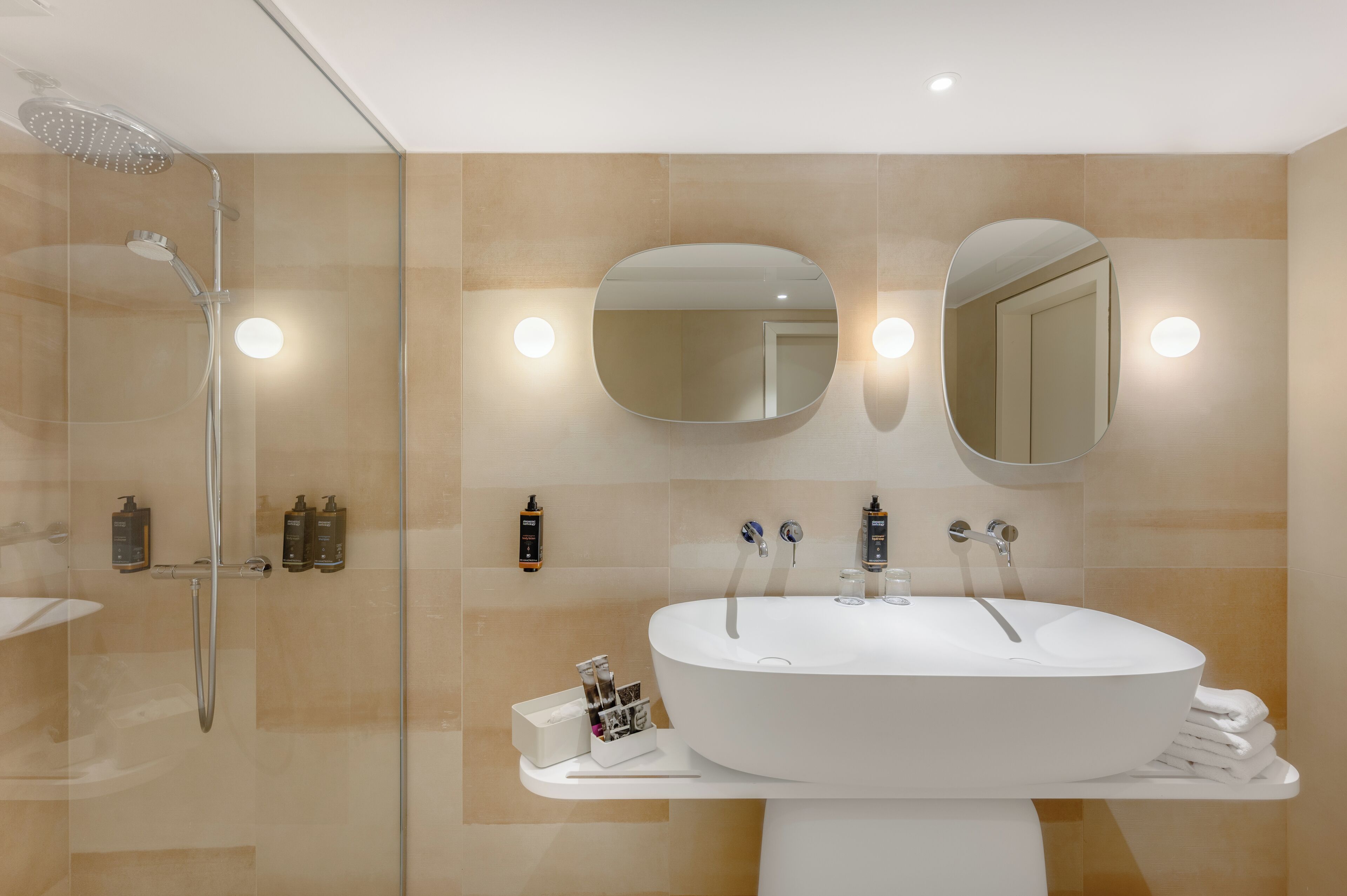 A modern bathroom with a large oval sink, two mirrors, a glass-enclosed shower, and toiletries on the wall.