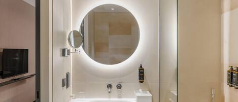 A modern bathroom with a round mirror, two sinks, a shower, and a television in the adjacent room.