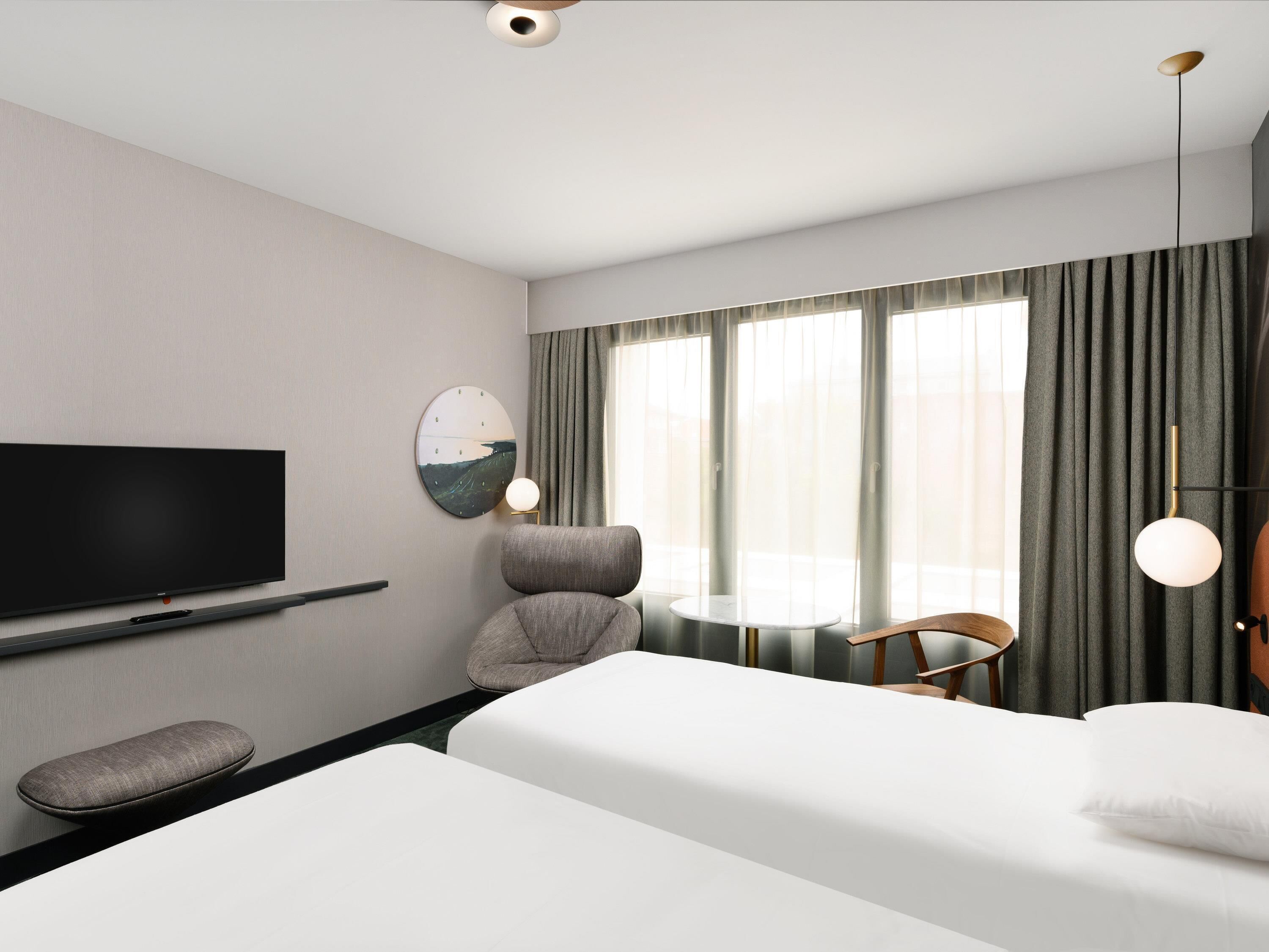 Una habitación de hotel moderna con una cama grande, un televisor de pantalla plana montado en la pared, una mesa redonda con una silla y un ventanal con cortinas.