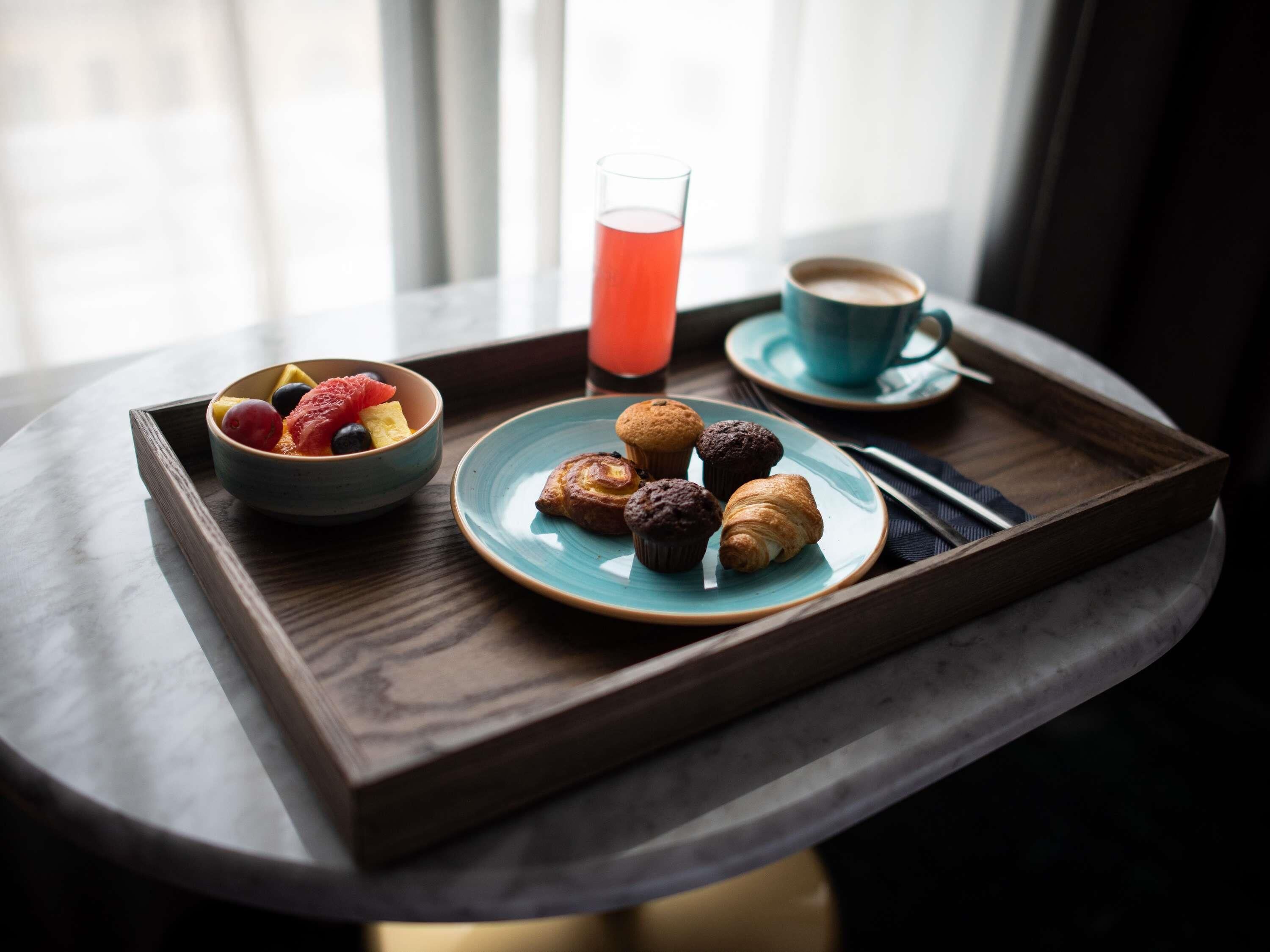 Uma bandeja com itens de café da manhã, incluindo croissants, muffins e uma tigela de frutas, além de uma xícara de café e um copo de suco.