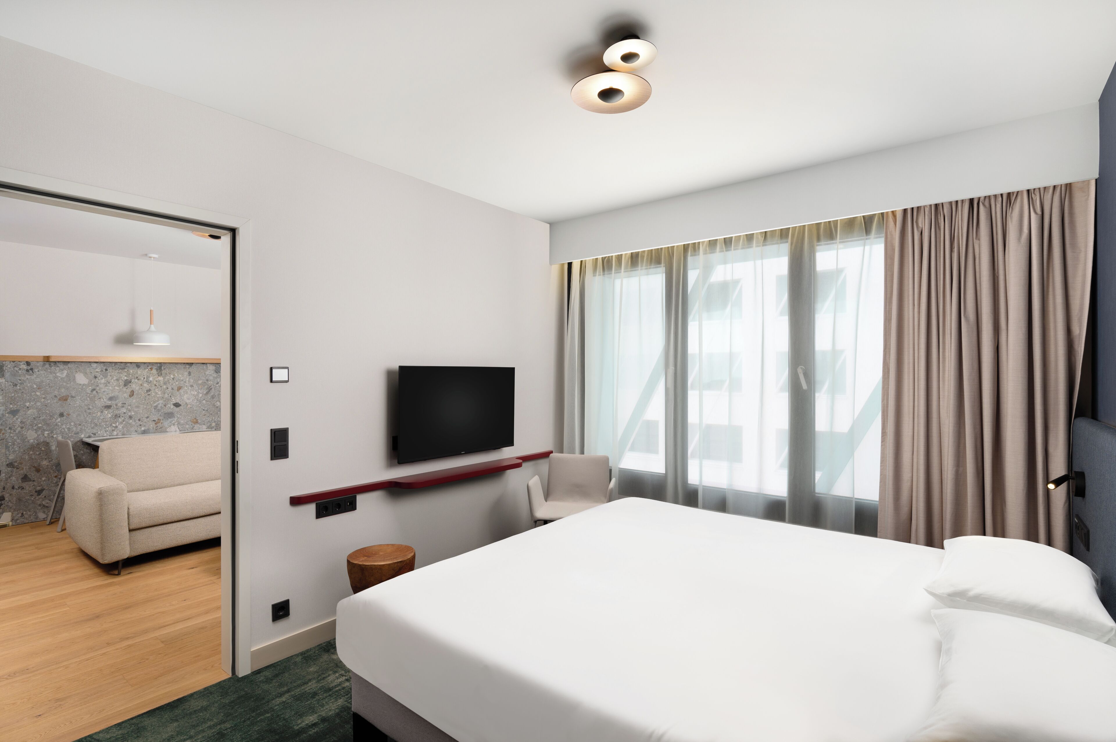 Ein modernes Hotelzimmer mit einem großen Bett, einem flachbildfernseher an der Wand, einer Sitzecke mit Sofa und Sessel sowie Blick auf die Stadt durch große Fenster.