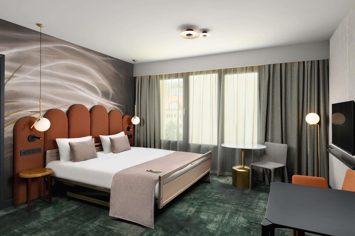 A hotel room with a bed, bedside tables, a desk, a chair, a TV, and a large window with curtains.