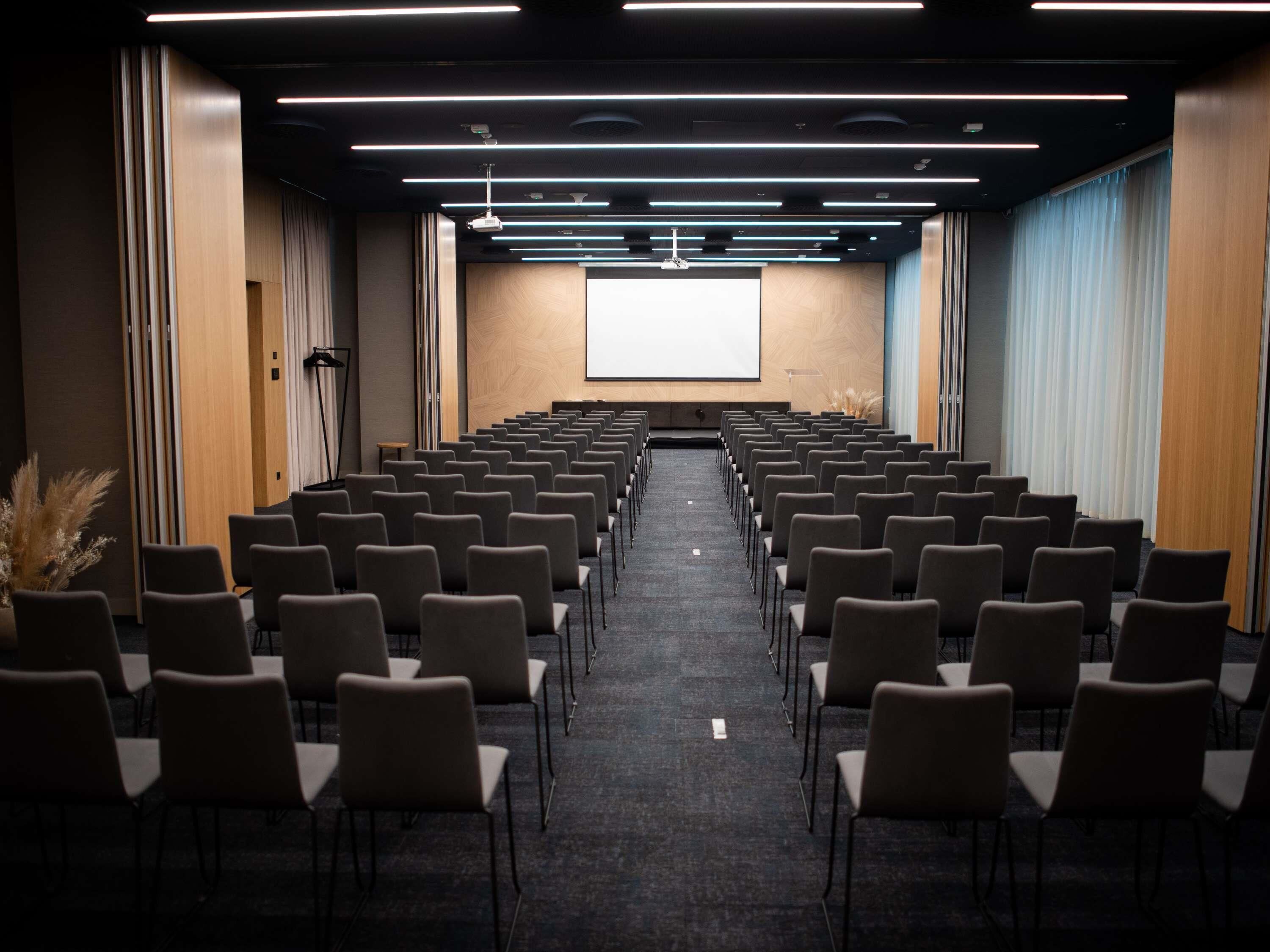 Un salón de conferencias con filas de sillas, una pantalla de proyector y suelo alfombrado.