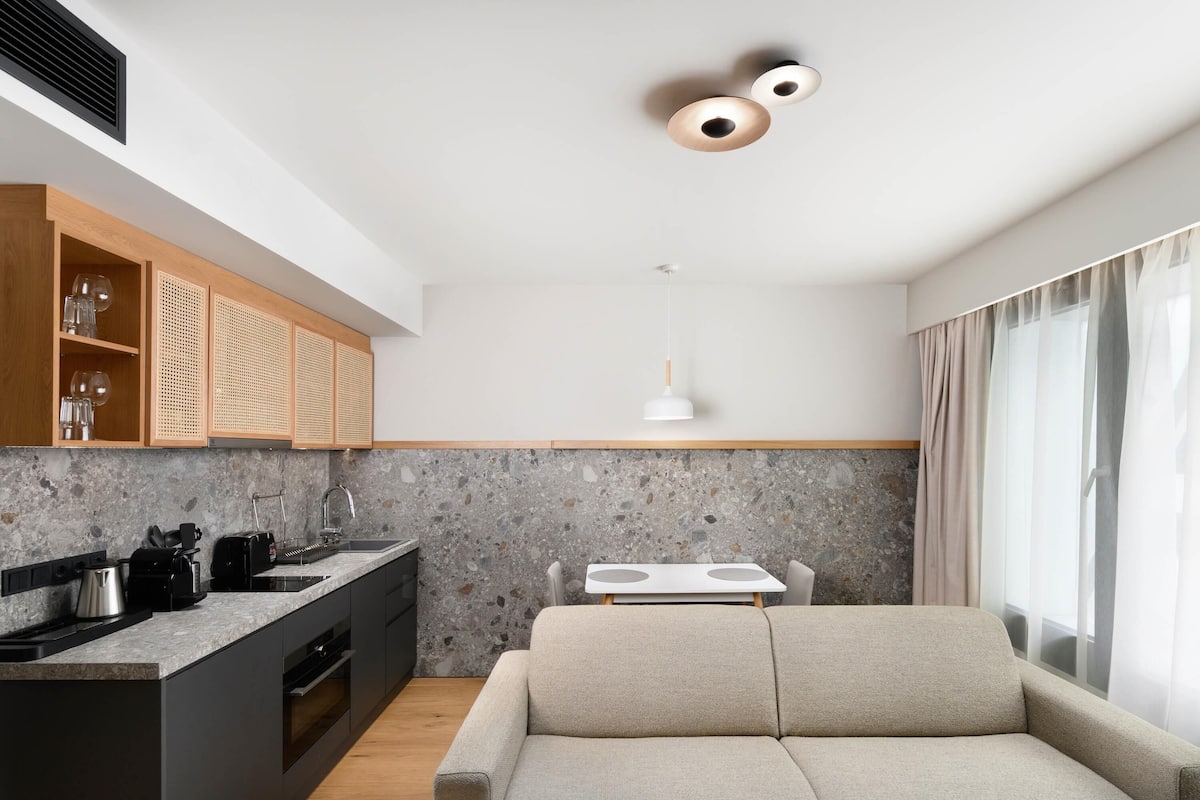A modern kitchen with a granite countertop, wooden cabinets, and a built-in oven. There is a dining area with a white table and a beige sofa.