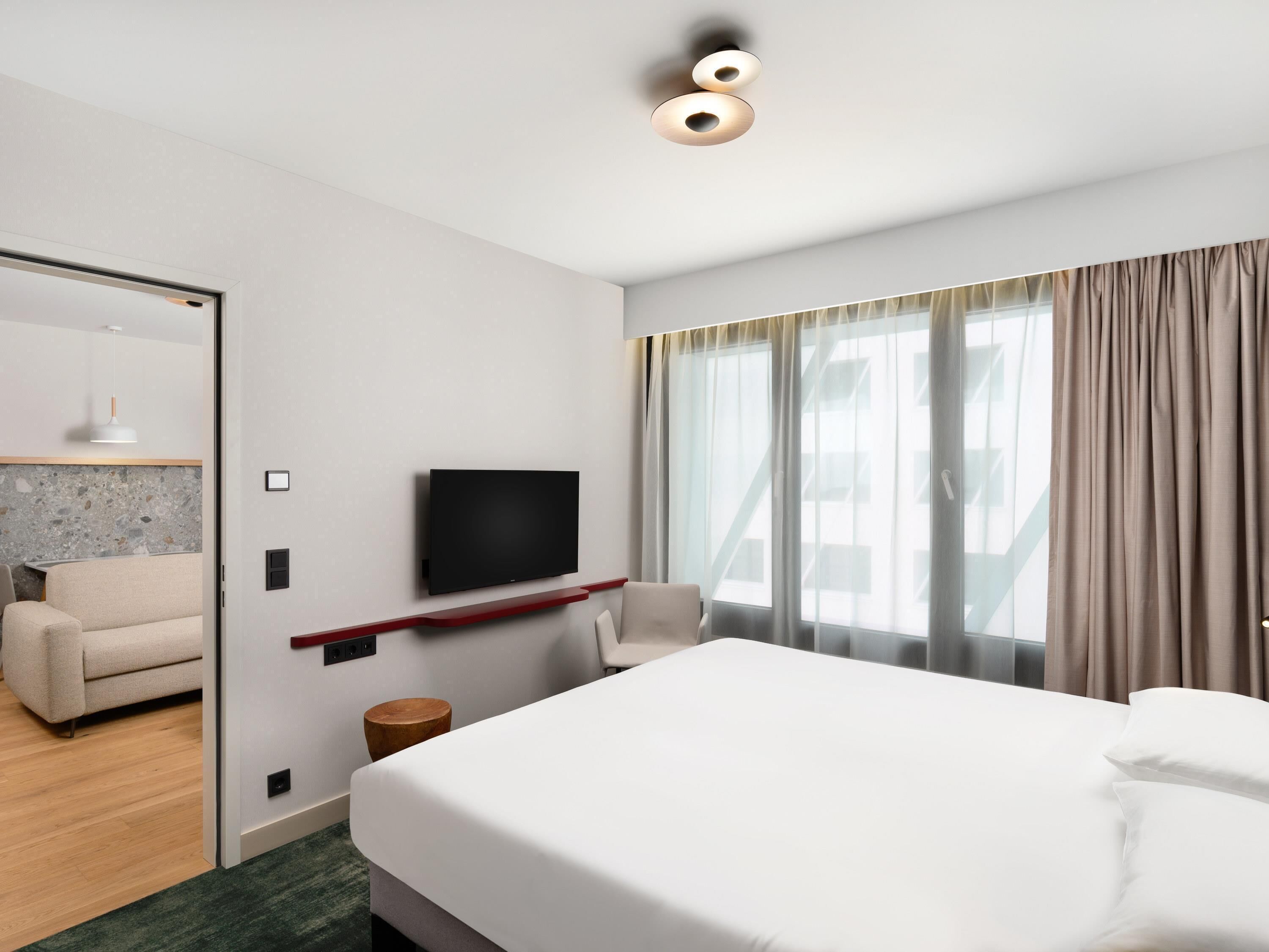 Een moderne hotelkamer met een bed, een aan de muur bevestigde televisie, een sofa en een raam met gordijnen.